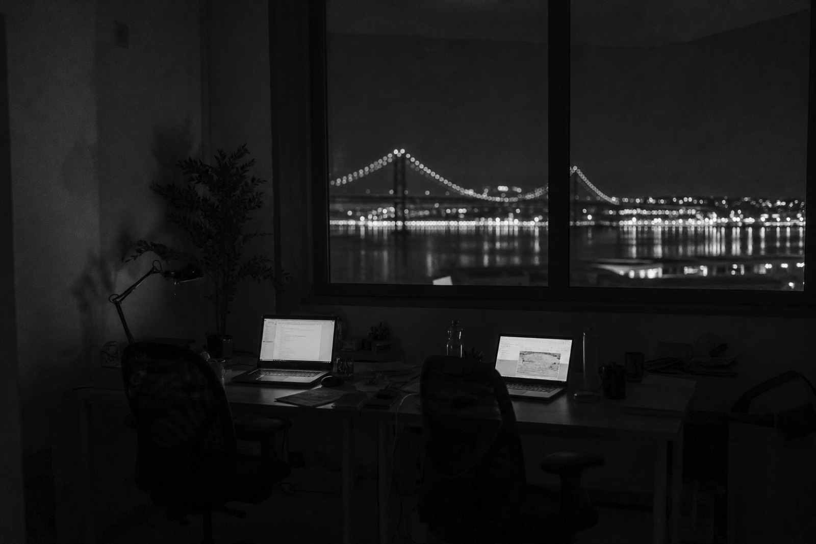 Two desks pushed together in an empty office at night, laptops open, the river behind the window